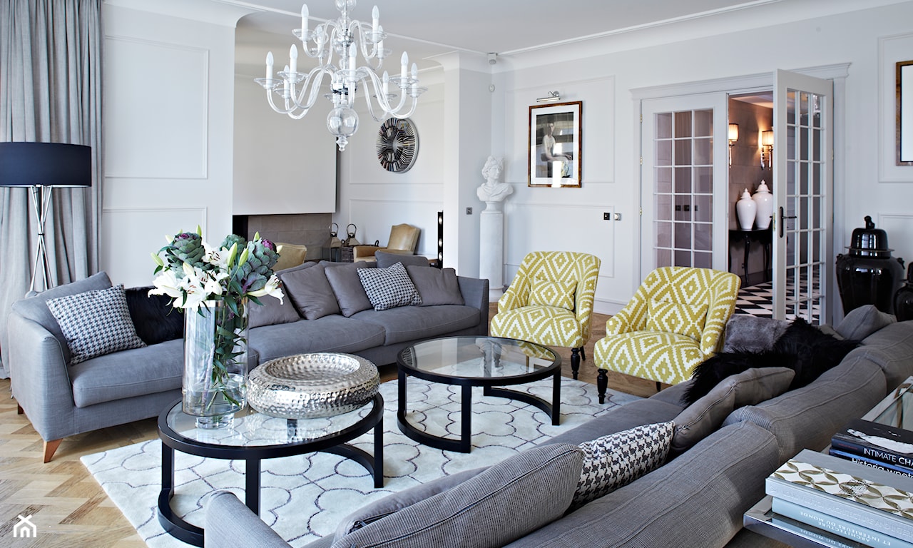 szara sofa, okrągły stolik ze szkła, żółte fotele w białe wzory, biały żyrandol, lampa podłogowa z czarnym abażurem