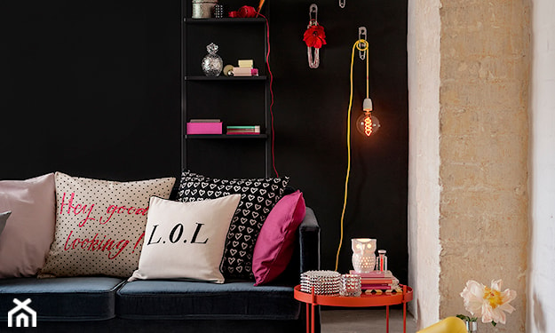 czarna ściana, metalowy stolik, lampa żarówka na żółtym kablu, różowe poduszki, beżowa cegła