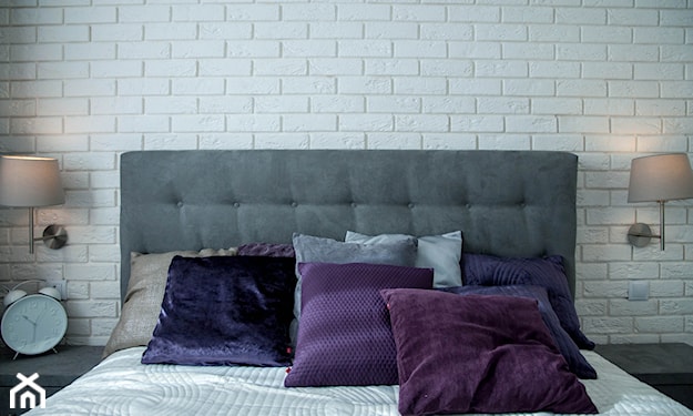 szare łóżko, biała cegła, fioletowe poduszki