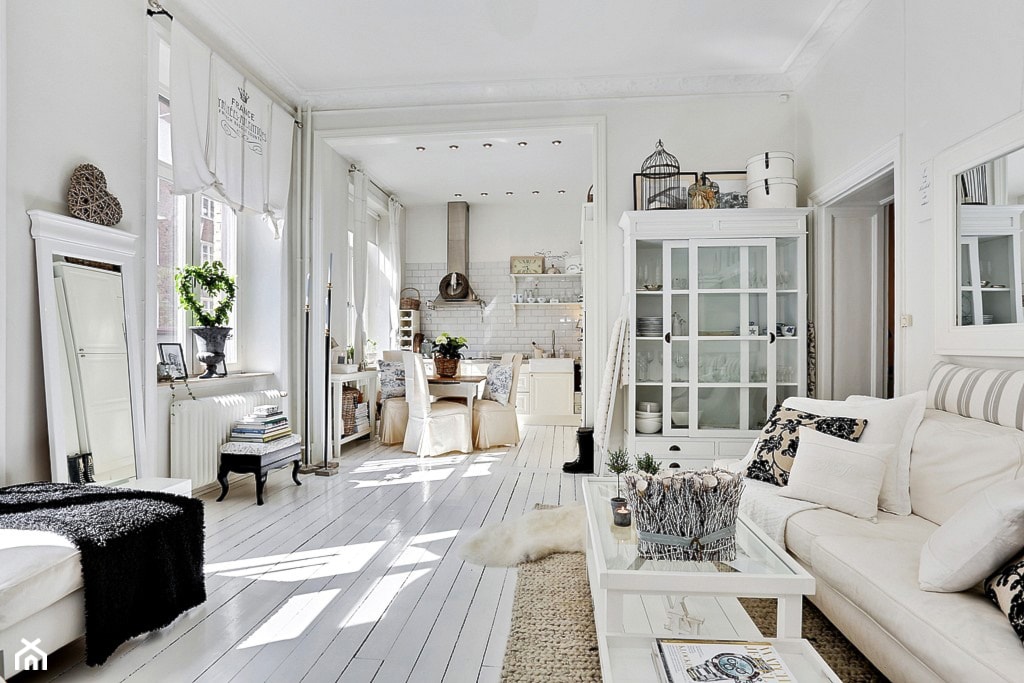 podłoga z białych desek, kremowa sofa, lustro w białej ozdobnej ramie, beżowy dywanik, czarna narzuta z długim włosiem