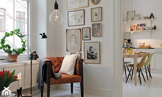 lampa wisząca - żarówka, białe ściany, brązowy pikowany fotel na nóżkach, retro obrazy w złotych ramach