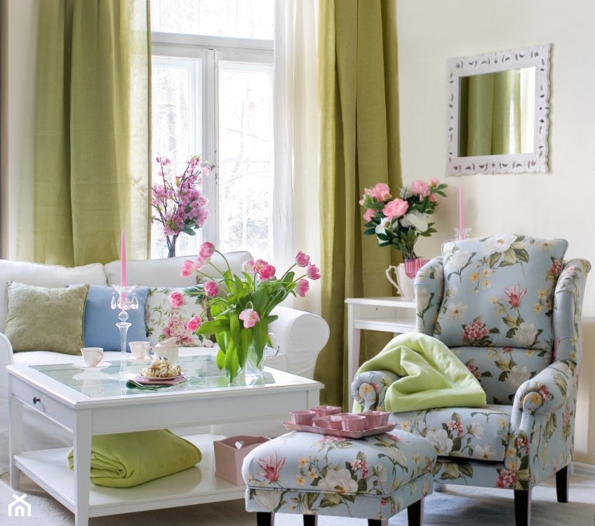 błękitny fotel w różowe kwiaty, biała sofa, długie zielone zasłony, biały stolik ze szklanym blatem