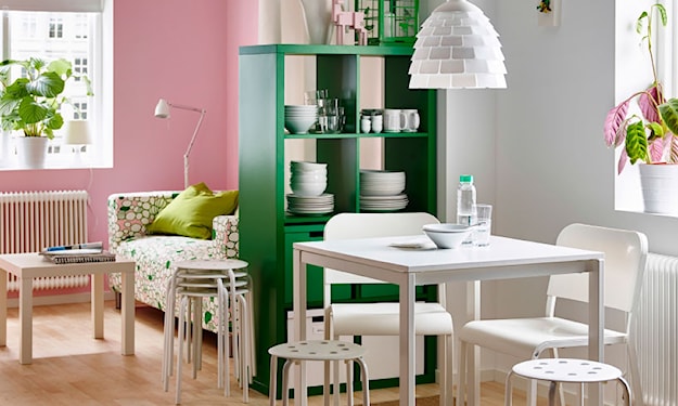 biały stół i taborety, lampa wisząca z białym abażurem, zielony regał, różowa ściana