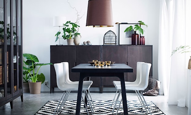 lampa z brązowym abażurem, czarny stół, białe krzesła na metalowych nogach, komoda z ciemnego drewna, biało-czarny dywan