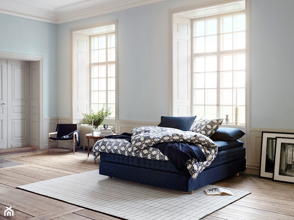 granatowe łóżko Hästens, drewniana podłoga, beżowy dywan, biała pościel w granatowe wzory, biała lamperia