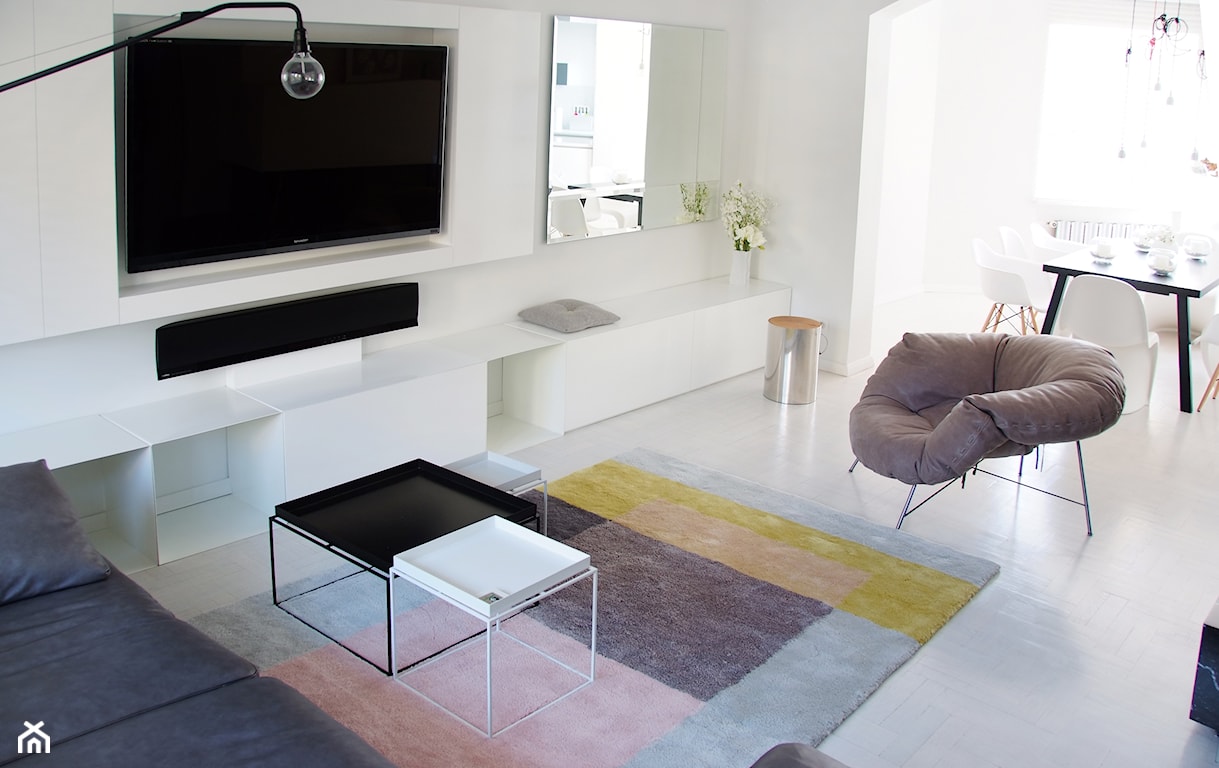 nowoczesny biały salon, dywan w kolorowe kwadraty, długa biała szafka rtv, lustro ścienne, szary fotel na metalowych nogach, meble z wgłębieniem na telewizor