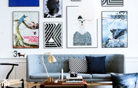 biała lamperia, szara sofa na drewnianych nóżkach, ściana zapełniona grafikami