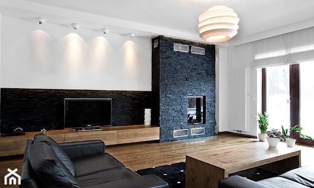 drewniana podłoga, czarna sofa ze skóry, czarna lamperia na ścianie, kominek na ścianie z kamienia ozdobnego