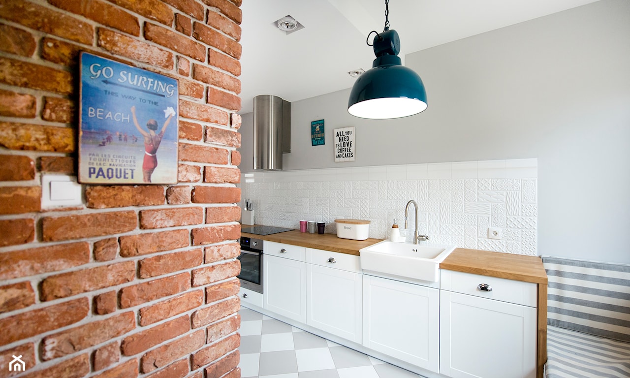ceglana ściana w kuchni, metalowa lampa, białe meble kuchenne z drewnianym blatem