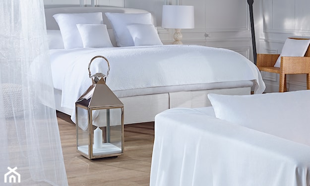 drewniana podłoga, białe łóżko, białe długie firany, metalowa latarenka ze świeczką