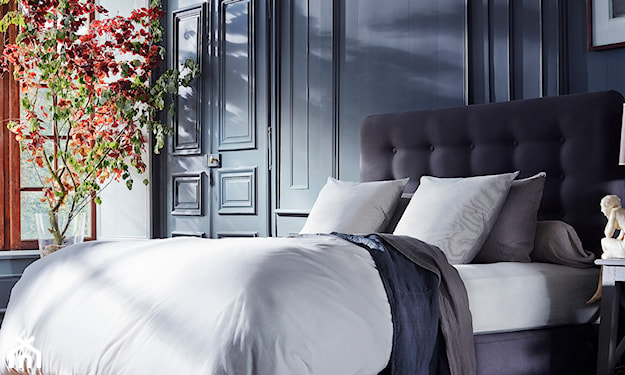 biała pościel, grafitowe łóżko z pikowanym zagłówkiem, drzewko ozdobne w sypialni