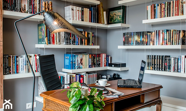 szare ściany, drewniane biurko z ozdobnymi nogami, metalowa lampa podłogowa, białe półki z książkami
